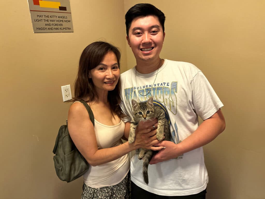 A woman and a young man stand together in a hallway, both smiling while holding a small gray tabby cat. The woman has long hair and is wearing a white top with a patterned skirt, while the man is in a graphic t-shirt and shorts.