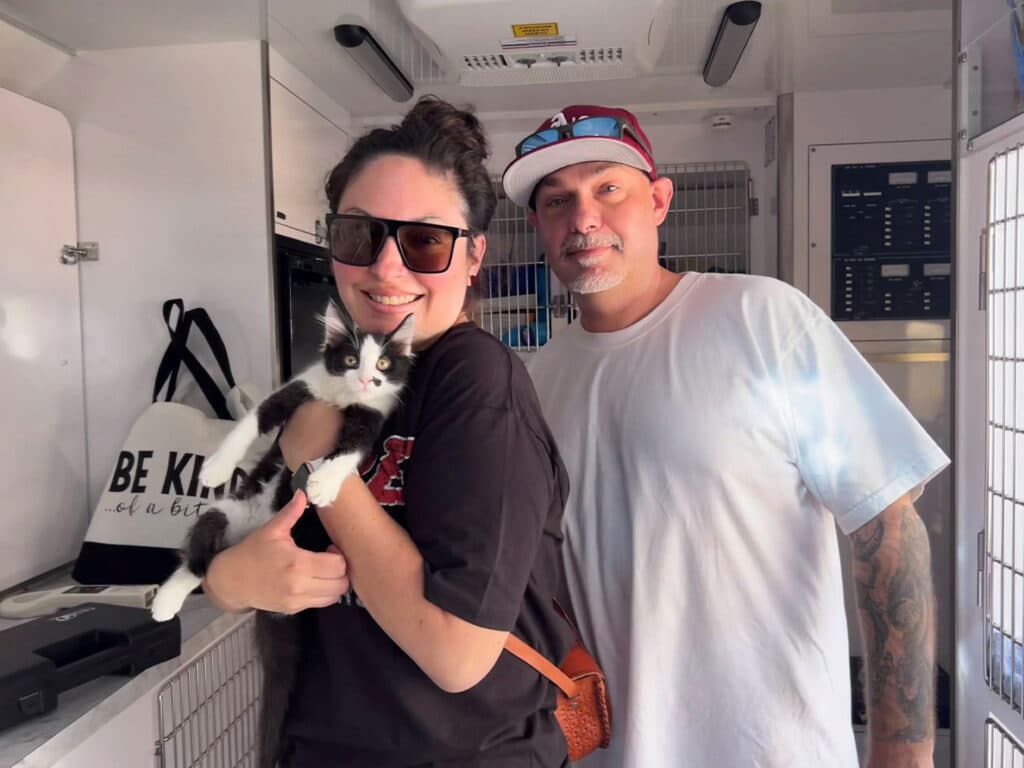 A woman wearing sunglasses holds a black and white cat in her arms, smiling. Beside her is a man with a shaved head and a tattooed arm, wearing a light blue t-shirt and a red cap. They are inside a mobile adoption vehicle.