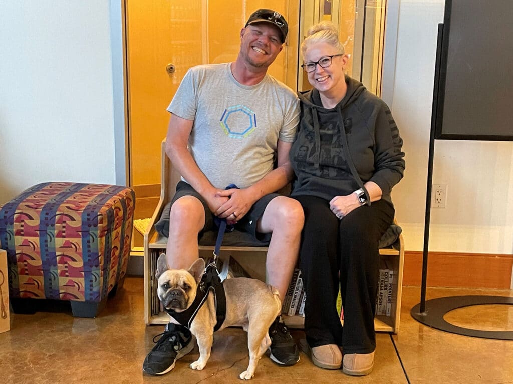 A man and woman sitting on a bench with a small dog on a leash in front of them. The man wears a gray t-shirt and shorts, while the woman is dressed in a dark hoodie and pants. A patterned chair is visible in the background.