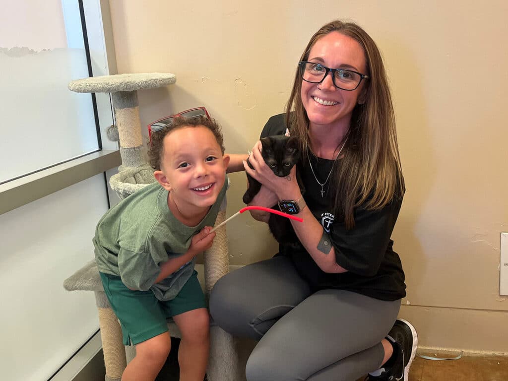 A young boy with curly hair smiles while standing next to a woman with glasses, who holds a small black kitten. They are seated next to a cat tree in an indoor space with natural light.