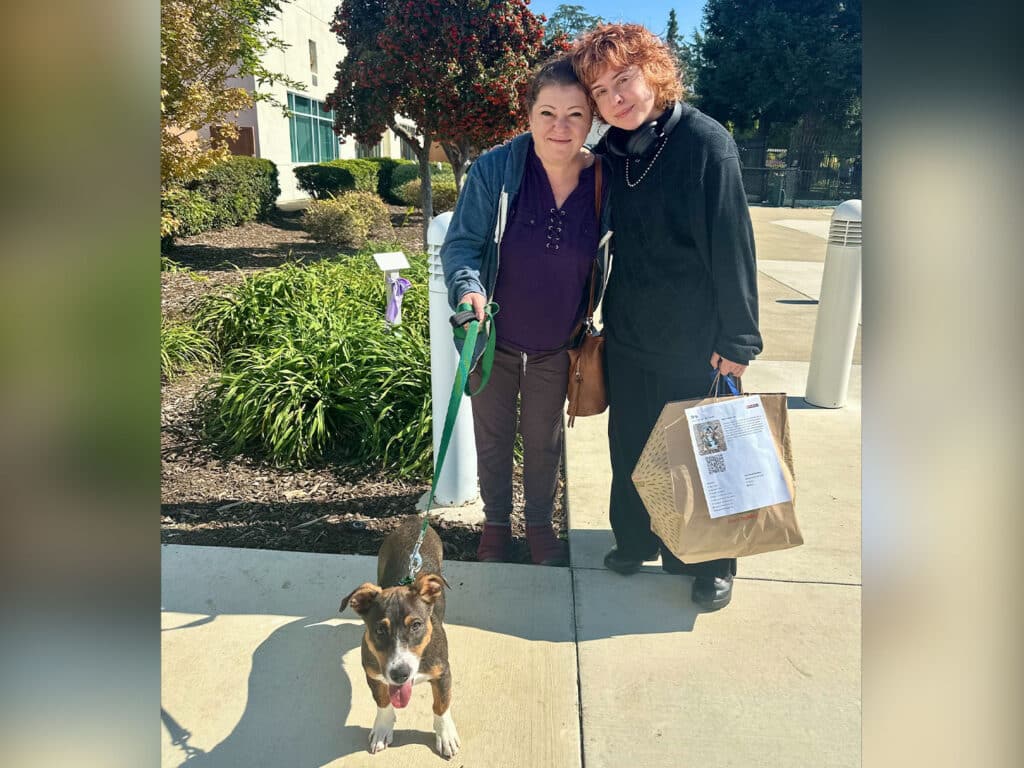 Two women stand together outside a building, smiling. One woman is holding a leash attached to a brown and white dog. The other woman is carrying a shopping bag. Green shrubs and trees are visible in the background under clear skies.