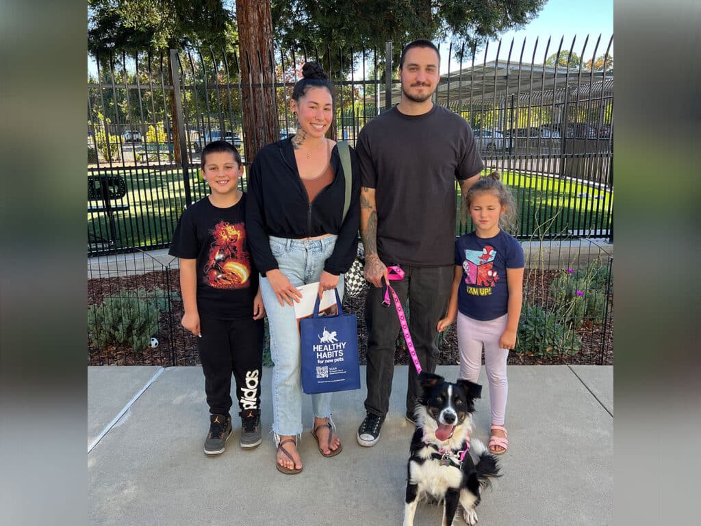 A family stands outside with a black and white dog on a leash. The group includes a woman wearing a black jacket, a man in a black shirt, a boy in a graphic tee, and a girl in a blue shirt, all smiling at the camera. Green trees and a fence are in the background.