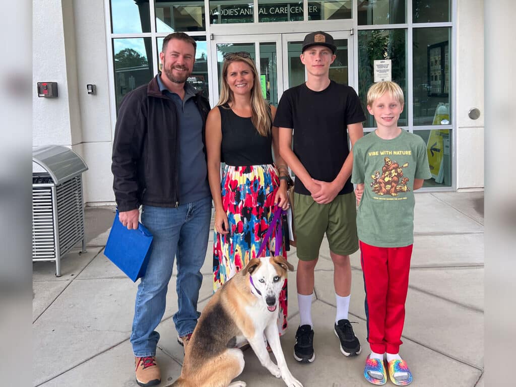 A family stands outside a building, smiling for the photo. The group includes a man in a jacket, a woman in a colorful dress, and two boys. A dog sits in front of them. The background features glass doors and a paved walkway.