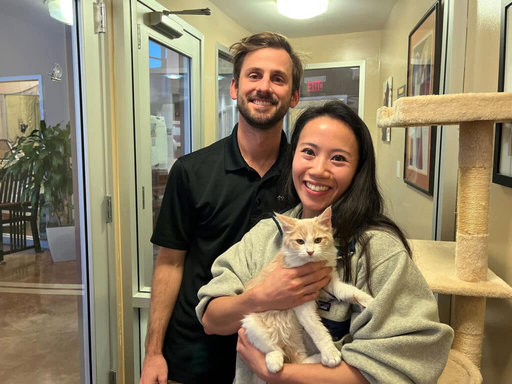 A man and a woman stand in a room with light-colored walls and a cat tree. The man, in a black polo shirt, smiles while the woman, wearing a gray sweater, holds a light-colored cat. They appear to be indoors, likely at an animal shelter.