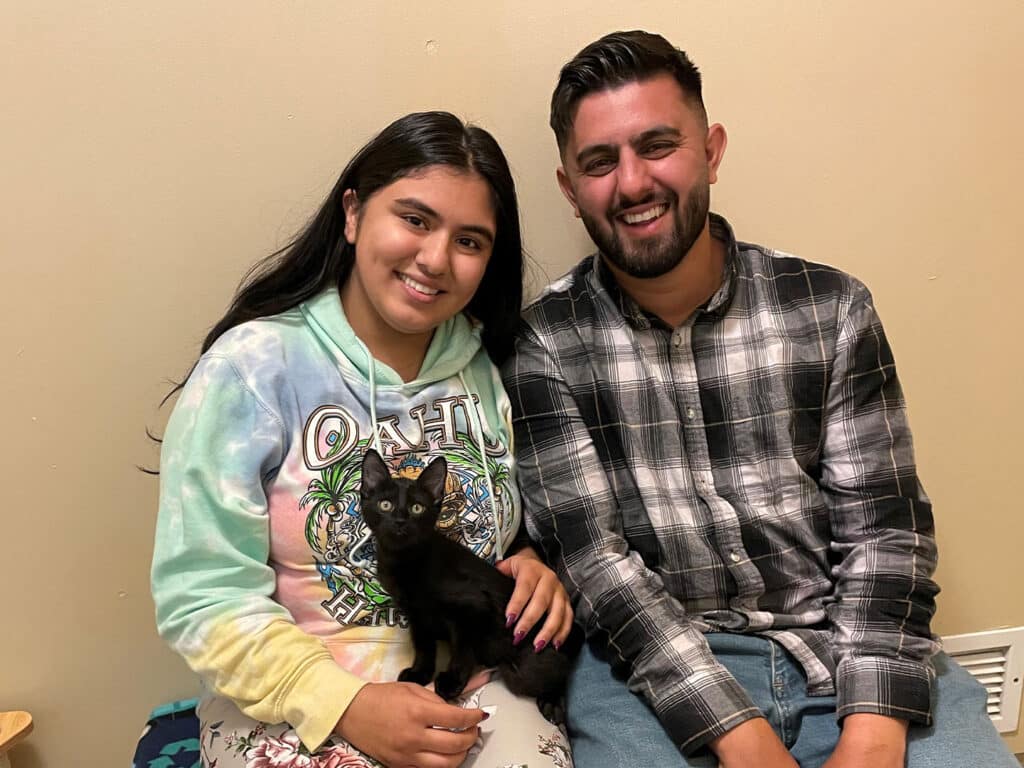 A young woman with long dark hair wearing a colorful hoodie sits on a couch, holding a black kitten. Beside her is a young man with short dark hair, smiling while wearing a plaid shirt. They appear relaxed and happy, with a neutral-colored wall in the background.