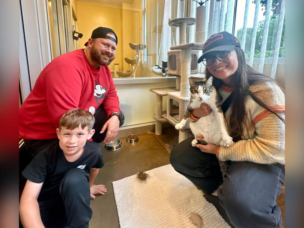 A man and a woman smile while holding a cat in a pet-friendly indoor space. A young boy sits on the floor nearby. Cat trees and toys are visible in the background, along with a bowl and a light-colored mat on the floor.