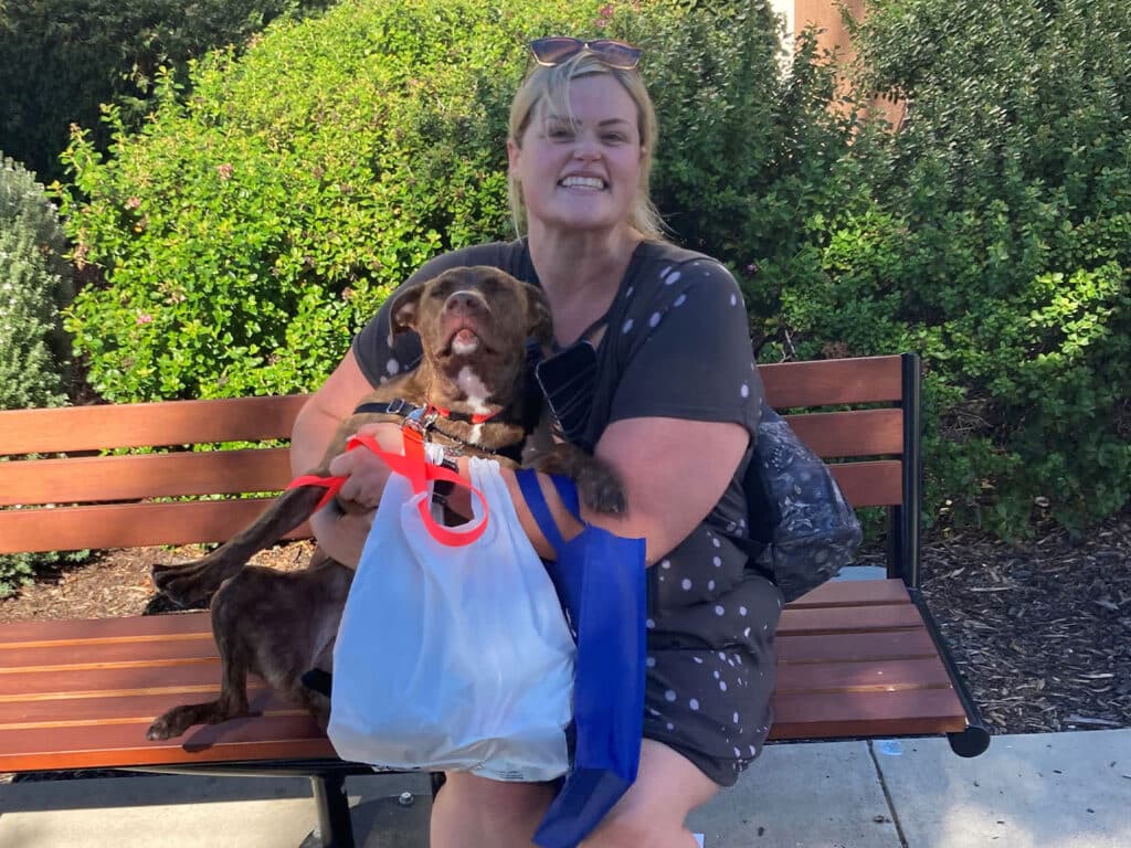 A woman with blonde hair, wearing a black dress with white polka dots, is sitting on a bench. She holds a brown dog in her lap and is smiling. A plastic bag is resting on her lap, and greenery is visible in the background.