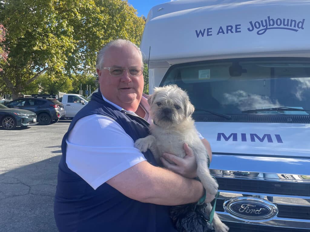 A man holding a small dog in front of a van that says "WE ARE Joybound". The man is wearing a dark vest over a white shirt, and the dog appears to be light-colored with a shaggy coat. Trees and vehicles are visible in the background.