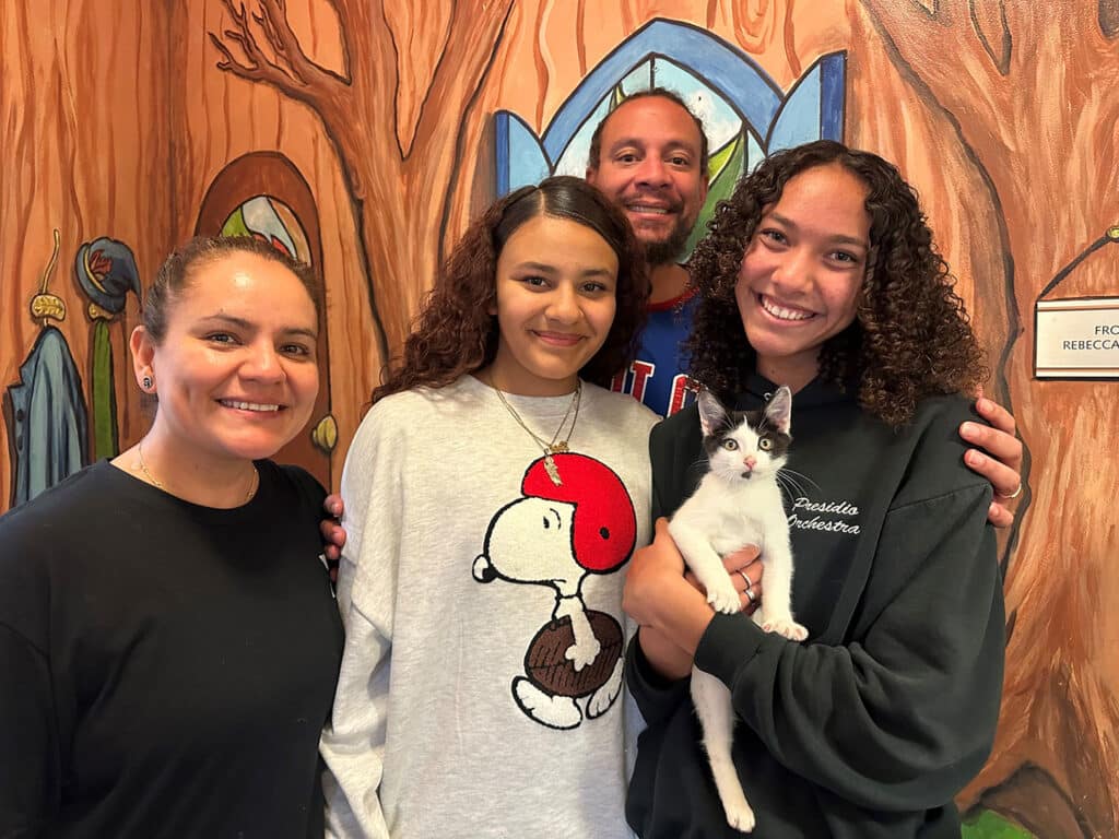 A group of four people poses together in front of a colorful mural. Two women stand on the left, one holding a cat. A young girl and a woman on the right hold a white cat with black markings. The background features a whimsical tree design.