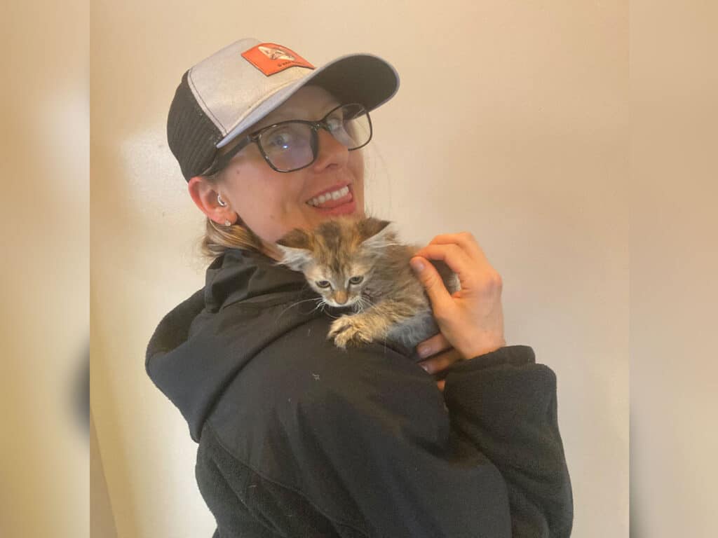 A woman wearing glasses and a baseball cap smiles while holding a small kitten against her shoulder. She is dressed in a black jacket, and the background is a light neutral color.