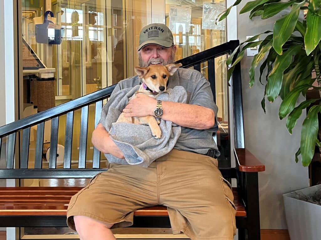 A man sitting on a wooden bench, holding a small dog wrapped in a grey blanket. He is wearing a cap and casual clothing. In the background, there is a stairway and indoor plants, creating a cozy atmosphere.