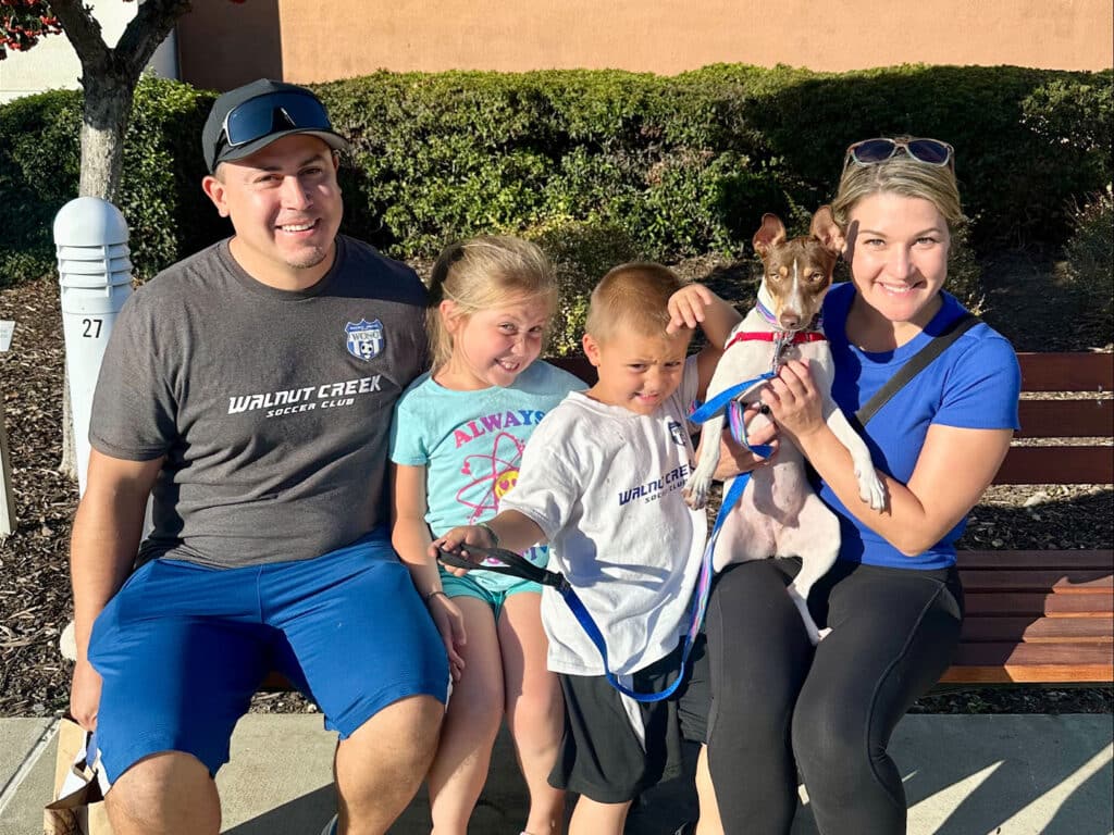 A family of four sits together on a bench: a man and woman, two children, one holding a small dog. The man wears a gray shirt and blue shorts, while the woman is in a blue shirt. The girl has a light blue shirt and the boy wears a white shirt.