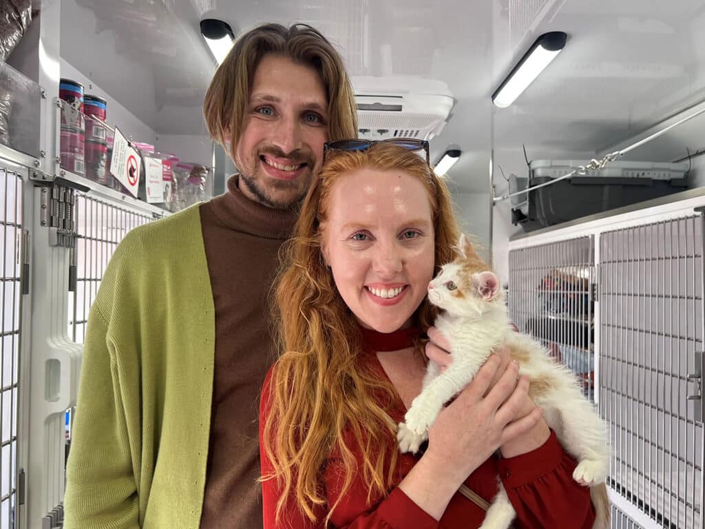 A man and a woman stand smiling together inside an animal shelter van. The woman holds a white and orange kitten in her arms. The background features several cages and supplies, indicating a pet adoption or care environment.