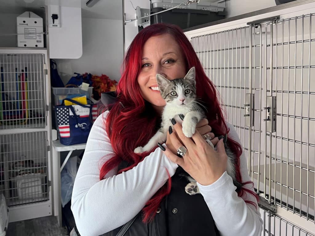 A woman with long red hair smiles while holding a gray and white kitten in her arms. She is wearing a black vest over a white long-sleeve shirt, and the background shows animal cages and supplies.