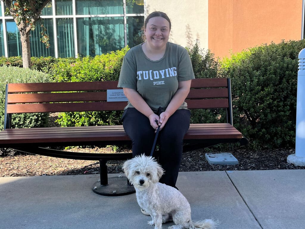 A woman sitting on a bench outdoors, wearing a "Studying" t-shirt, smiles while holding a leash connected to a small, fluffy white dog sitting beside her. Green shrubs and a building are visible in the background.