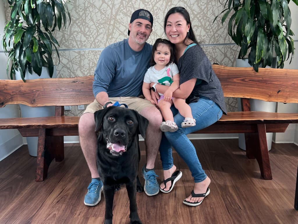 A man and a woman sit on a wooden bench, holding a toddler between them. The woman is smiling, while the girl wears a light shirt and looks at the camera. A black dog sits in front of them, looking directly at the viewer. Green plants are in the background.