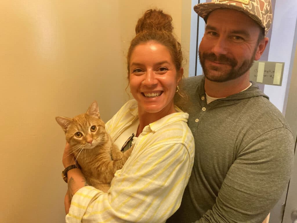 A woman with long brown hair in a white shirt holds an orange tabby cat, smiling. Next to her, a man with a beard wearing a gray shirt and a patterned cap stands, also smiling, in a well-lit hallway.