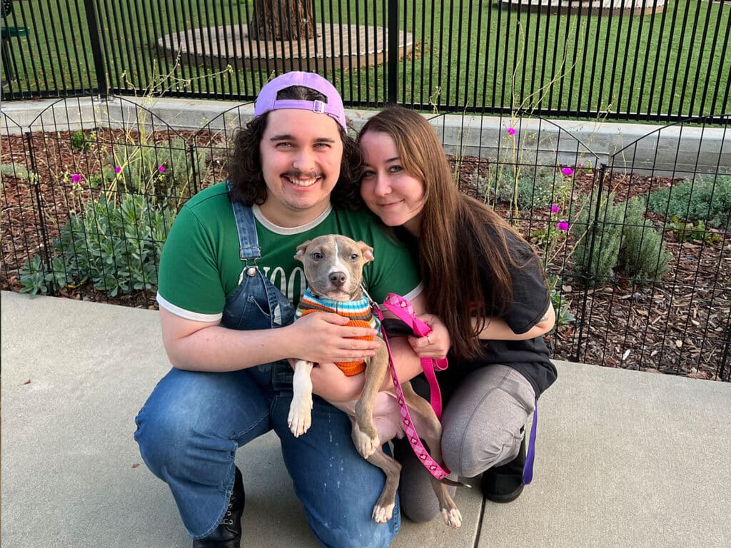 A man and a woman smiling while sitting on a sidewalk, holding a small dog. The man is wearing a green shirt with overalls and a purple hat, while the woman has long brown hair and is dressed in a black shirt and gray pants. They are surrounded by greenery and purple flowers.