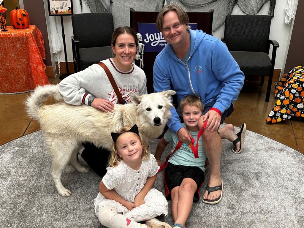 A family of four poses together in a room decorated with Halloween themes. Two children, a boy and a girl, are sitting on the floor with a white dog. The parents, a man and a woman, are smiling nearby. The scene has a warm and engaging atmosphere.