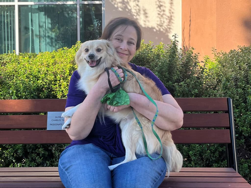 A woman with brown hair, wearing a purple shirt and blue jeans, sits on a wooden bench holding a small, smiling dog. The dog has light fur and is wearing a green harness. In the background, there are green bushes and a large window.
