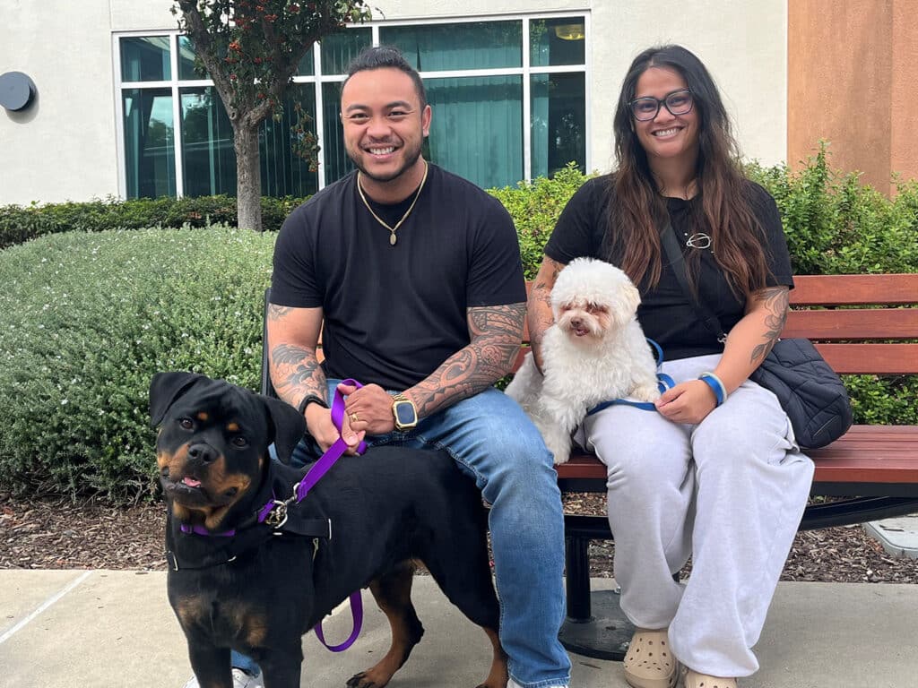A man and a woman sit on a bench outdoors, both smiling. The man, with tattoos and wearing a black shirt, has a Rottweiler beside him on a leash. The woman, wearing comfortable clothing, holds a small white dog on her lap.