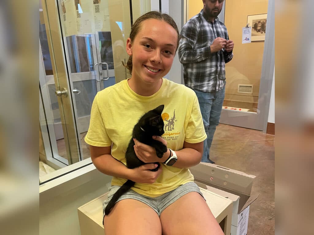 A young woman wearing a yellow t-shirt sits on a box holding a black kitten. In the background, a man in a plaid shirt stands, looking at his phone near a glass enclosure.