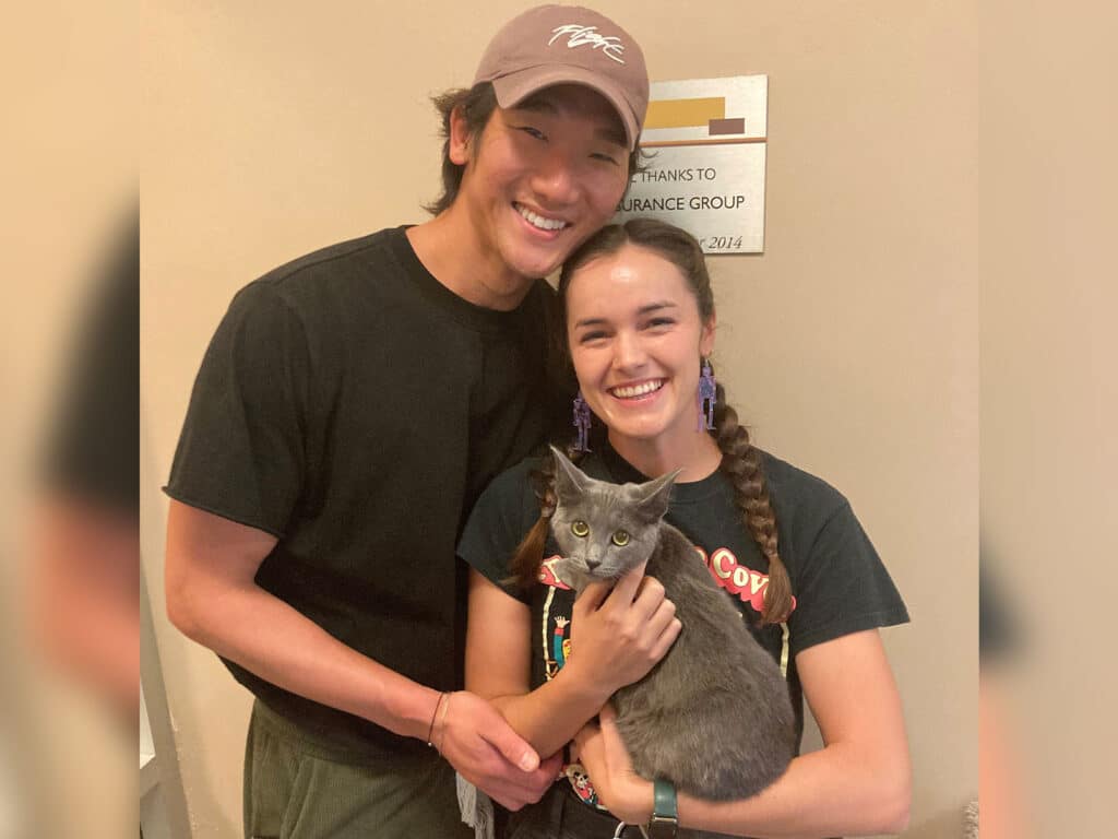 A young man in a black t-shirt and a baseball cap stands next to a young woman in a black shirt. She holds a gray cat in her arms, both smiling at the camera. A thank-you sign is visible in the background.
