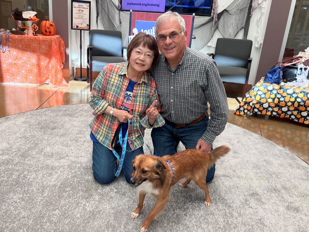 A woman and a man kneel on a gray rug next to a small brown dog. The woman holds the dog's leash and wears a plaid jacket. The man, dressed in a gray shirt, smiles at the camera. In the background, there are decorations, including orange and black colors.