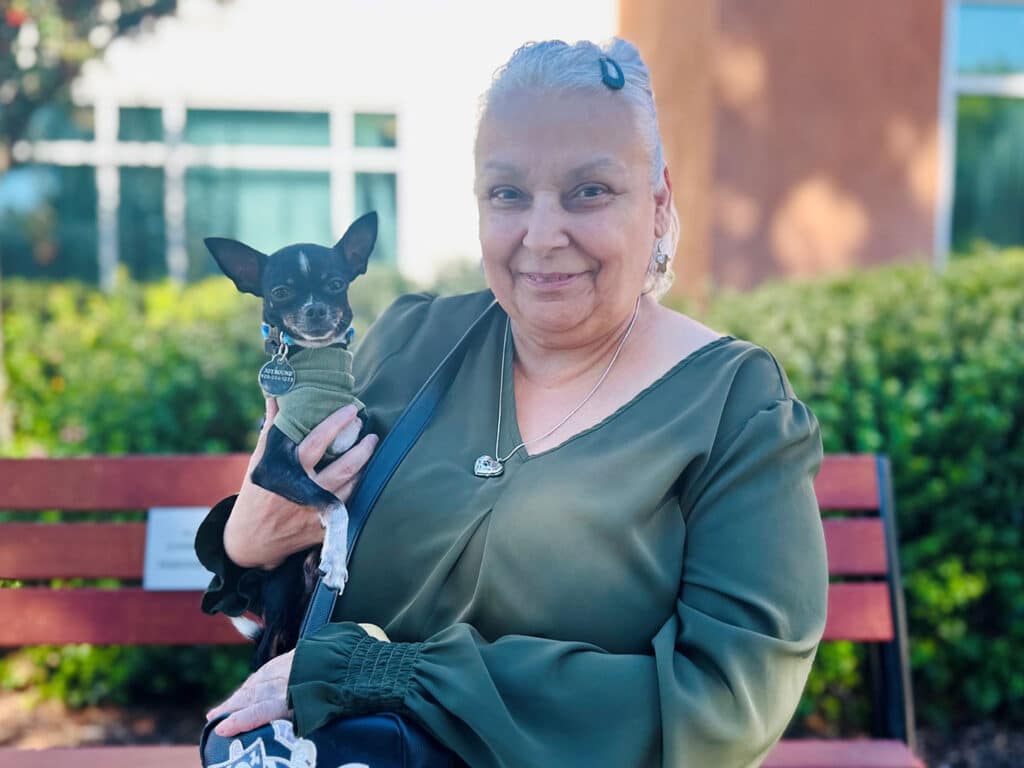 A woman with gray hair, wearing a green blouse, holds a small black dog on her lap while sitting on a wooden bench in a garden. The background features green foliage and a building. Both appear relaxed and content.
