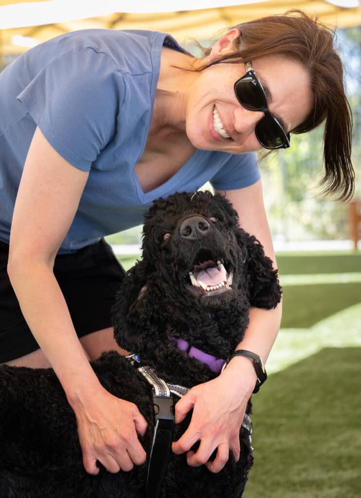 A woman wearing sunglasses and a blue shirt smiles while kneeling beside a black dog. The dog appears happy, showing its tongue and has a purple collar. They are on green grass under a canopy, suggesting a sunny outdoor setting.