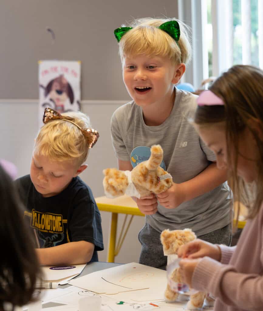 A boy with blonde hair wearing green cat ear headbands smiles while holding a plush toy. Nearby, a girl in a pink headband focuses on a craft project, and another boy in a black shirt draws at a table filled with art supplies.