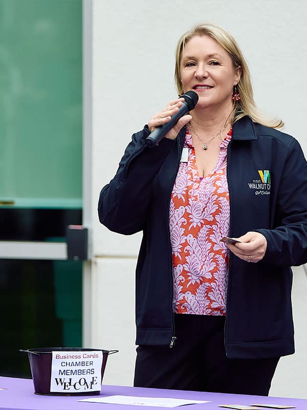 A woman speaking into a microphone at an event, wearing a navy jacket with the text 'Walnut Creek Chamber of Commerce'. She holds a business card and stands next to a bowl labeled 'Business Cards Chamber Members Welcome'. The setting suggests a community gathering, relevant for Joybound Rentals.