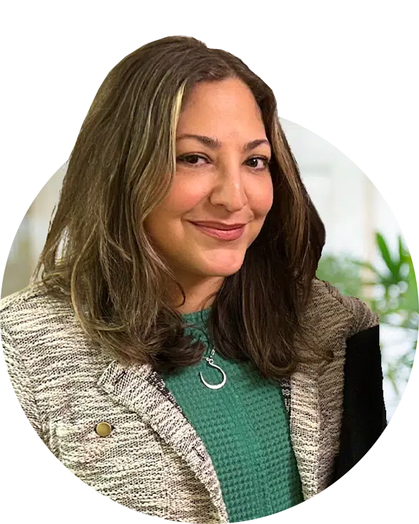 A woman with long, wavy brown hair, wearing a green sweater and a patterned blazer, smiles while standing against a blurred indoor background with greenery. The image relates to Joybound donate items, highlighting community support in giving societies.