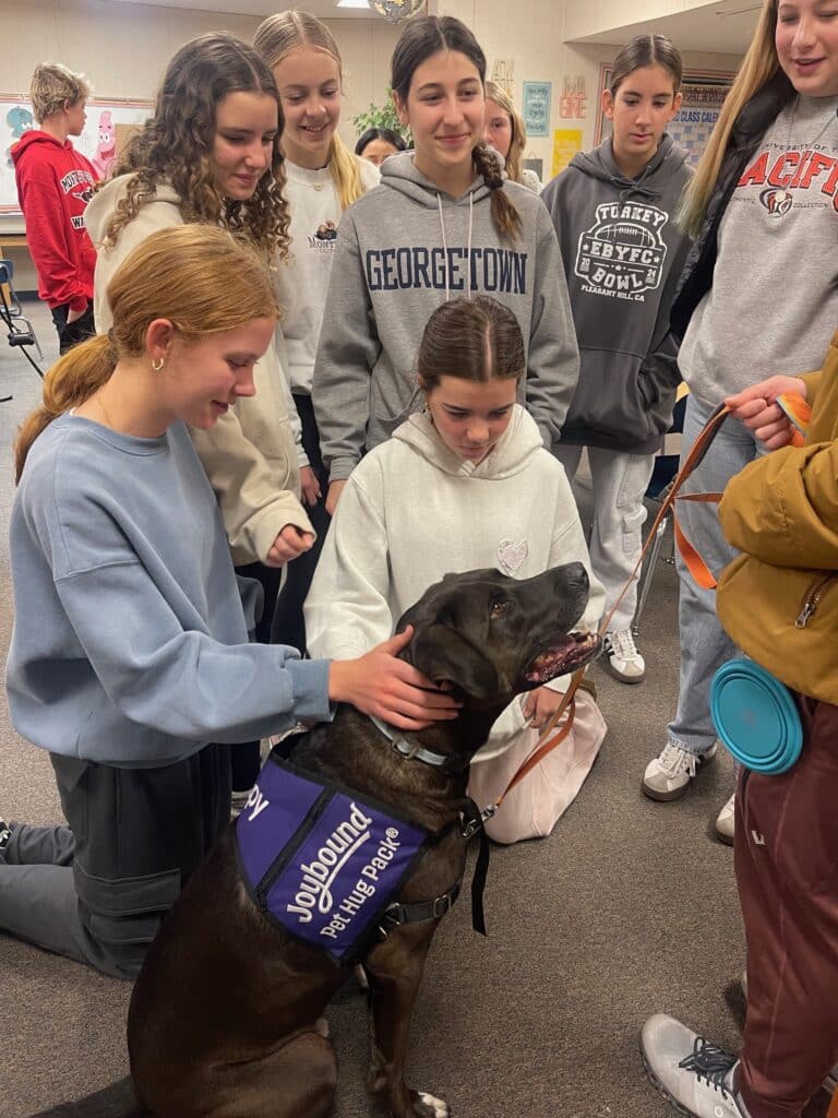 A group of teenage girls gathers around a therapy dog wearing a vest labeled "Jougbound - Helping People." One girl kneels to pet the dog, while others watch and interact, all within a bright classroom setting.