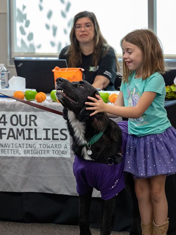 A young girl in a turquoise shirt and purple skirt pets a large black dog wearing a purple shirt. In the background, a woman sits behind a table with fruit and materials related to Joybound Rentals, which promotes family activities.