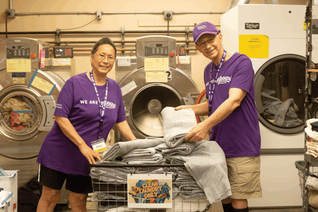 Two volunteers in matching purple shirts stand beside a cart filled with neatly stacked towels in a laundromat. Laundry machines are visible in the background, with yellow and blue labels, creating a supportive environment for community service.