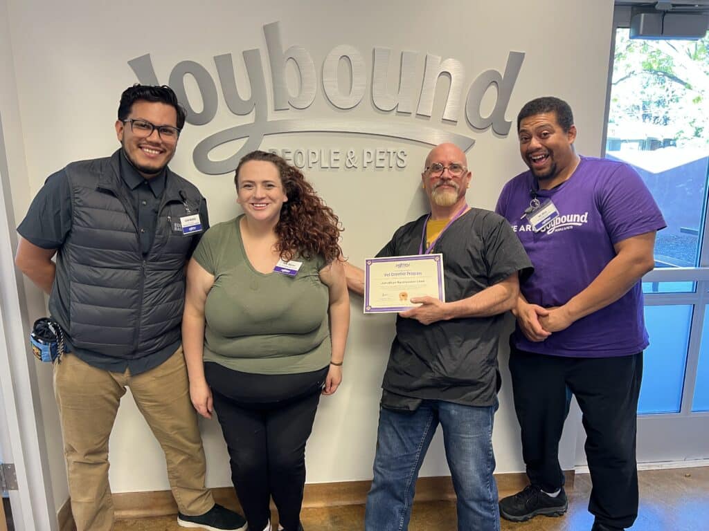 Four people stand in front of a Joybound sign. A man in scrubs holds a certificate while smiling. Two women, one in a green top and another in a purple shirt, smile next to him. A man in a vest stands on the left, all appearing to celebrate an achievement.