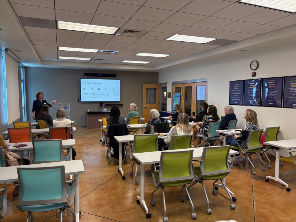 A classroom setting with a presenter speaking to an audience seated at colorful desks. A screen displays information, and various charts are on the walls. The space is well-lit, showcasing modern facility rentals available through Joybound Rentals.