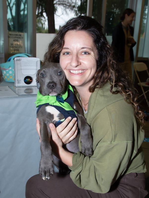 A woman with curly hair smiles while holding a gray puppy dressed in a green and navy outfit. The setting appears to be a rental facility, with chairs and decorations visible in the background. This image is associated with Joybound Rentals.