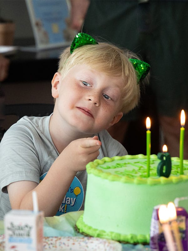 A young boy wearing green cat ears gazes at a vibrant green birthday cake topped with candles. The cake has a number 8 decoration. A small dessert container is visible on the table. This image relates to Joybound Rentals, showcasing a festive atmosphere.