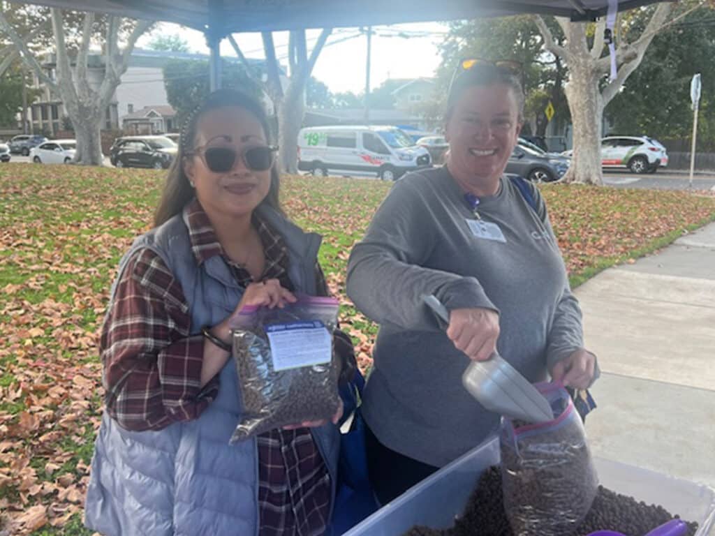 Two women are standing at a table outdoors under a tent. One woman, wearing sunglasses and a plaid shirt, holds a bag of food, while the other woman, in a gray sweatshirt, uses a scoop to fill a bag. The ground is covered with fallen leaves.