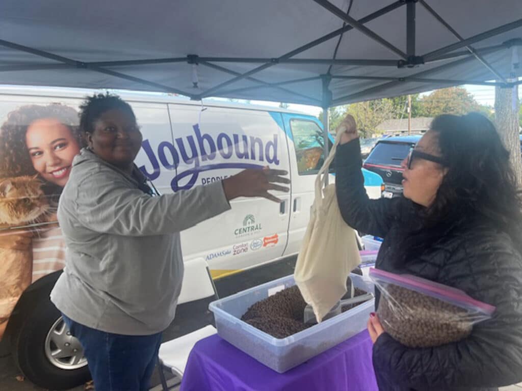 Two women are participating in a food distribution event under a tent. One woman hands a fabric bag to the other, who is holding a container of food. In the background, a van with a promotional design is visible.