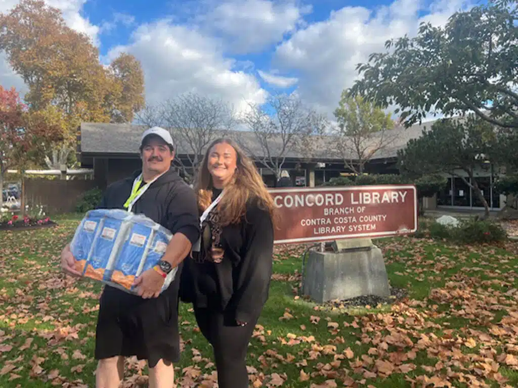 A man holding a box of food stands next to a woman outside the Concord Library, with autumn leaves on the ground and a cloudy sky in the background. The library's sign is visible behind them.
