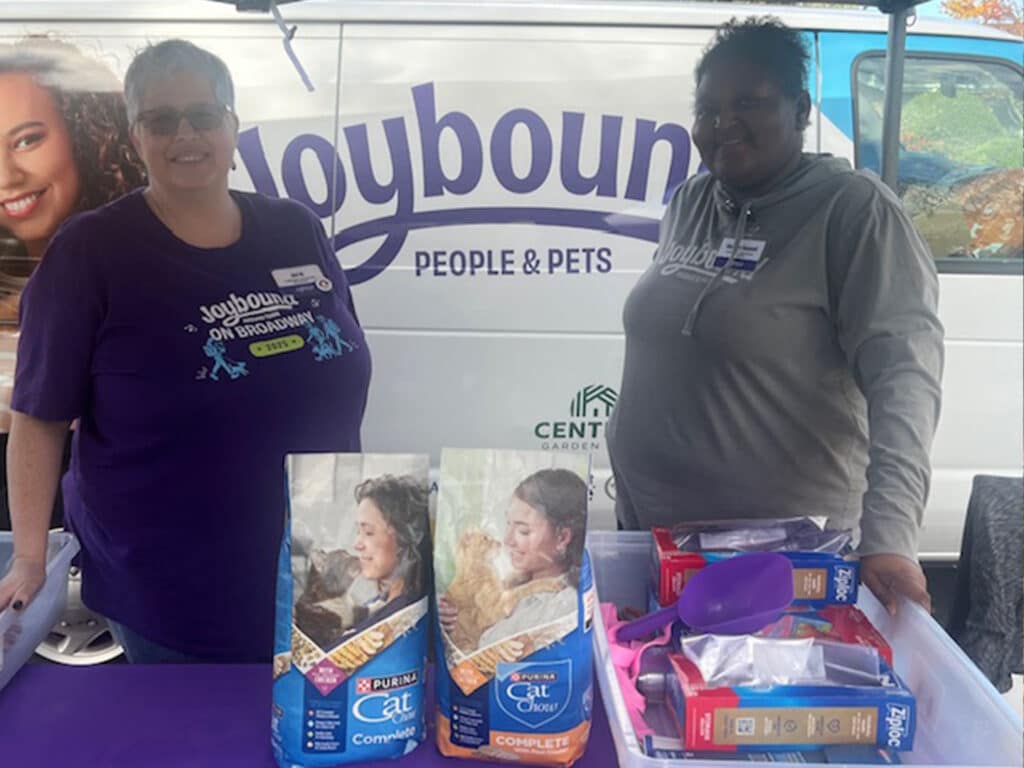 Two women standing behind a table with various pet food bags and supplies. One woman, wearing a purple shirt, smiles at the camera, while the other wears a gray sweatshirt. A colorful van with the text 'Joybound People & Pets' is in the background.
