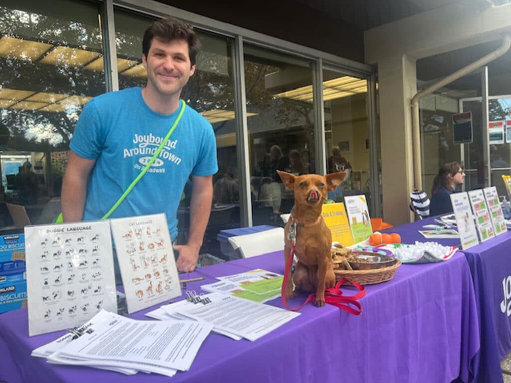 A man wearing a blue shirt stands behind a table covered with pamphlets and information on animal care. Next to him is a small dog sitting calmly, both appearing welcoming in a community event setting.