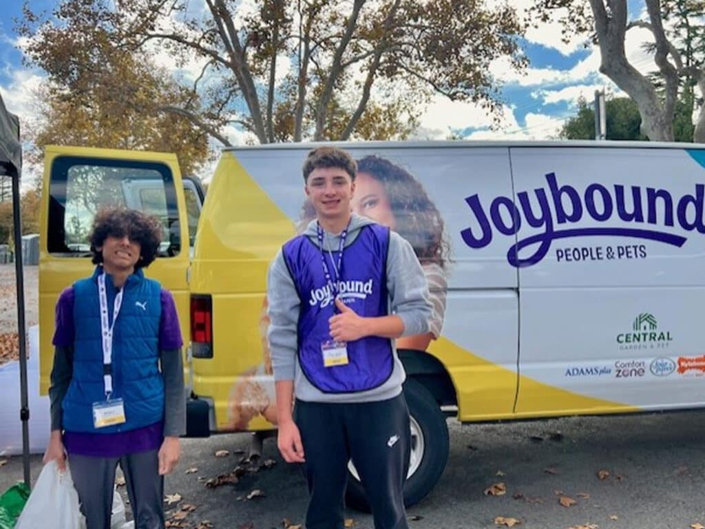 Two young volunteers stand in front of a van labeled 'Joybound People & Pets.' One wears a purple vest with the logo, while the other wears a blue jacket. The background includes trees and a partly cloudy sky, creating a community event atmosphere.