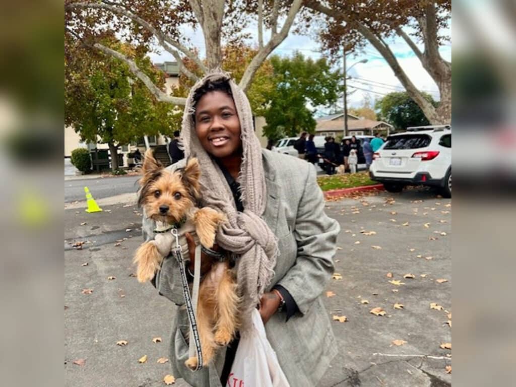 A woman wearing a gray coat and a scarf holds a small dog in her arms, smiling at the camera. In the background, people are seen behind parked cars under trees with autumn leaves.