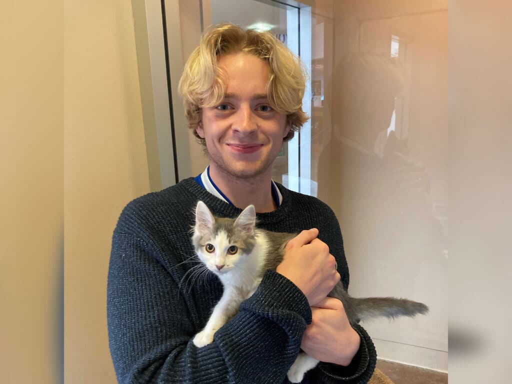 A young man with wavy blond hair smiles while holding a gray and white kitten in his arms. He is wearing a dark sweater and standing in a brightly lit interior near a glass door.