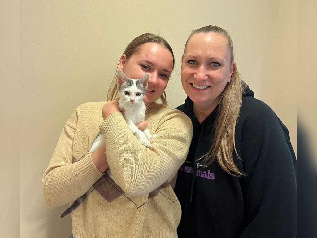 Two women stand together, smiling. One woman holds a small white kitten, while the other stands beside her. They are posed against a light-colored wall, and both are wearing casual clothing.