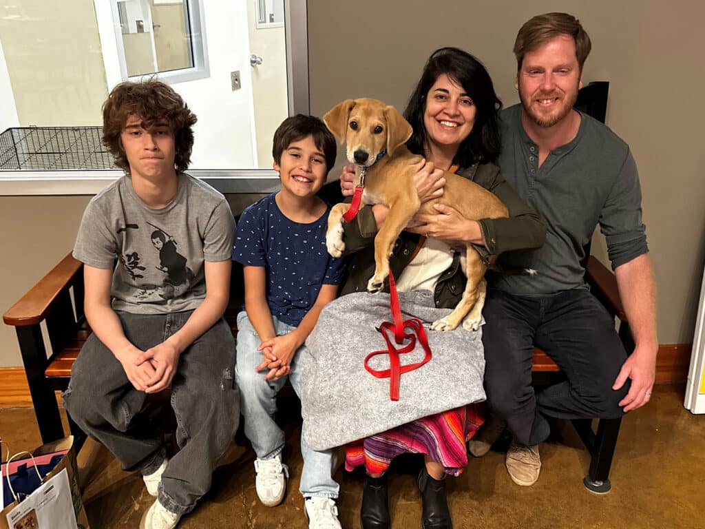 A family of four sits together on a bench in a room. The mother holds a small dog with a red leash, while the father sits next to her. Two boys sit on the left, one wearing a gray shirt and the other in a blue shirt. A welcoming, casual environment is visible.